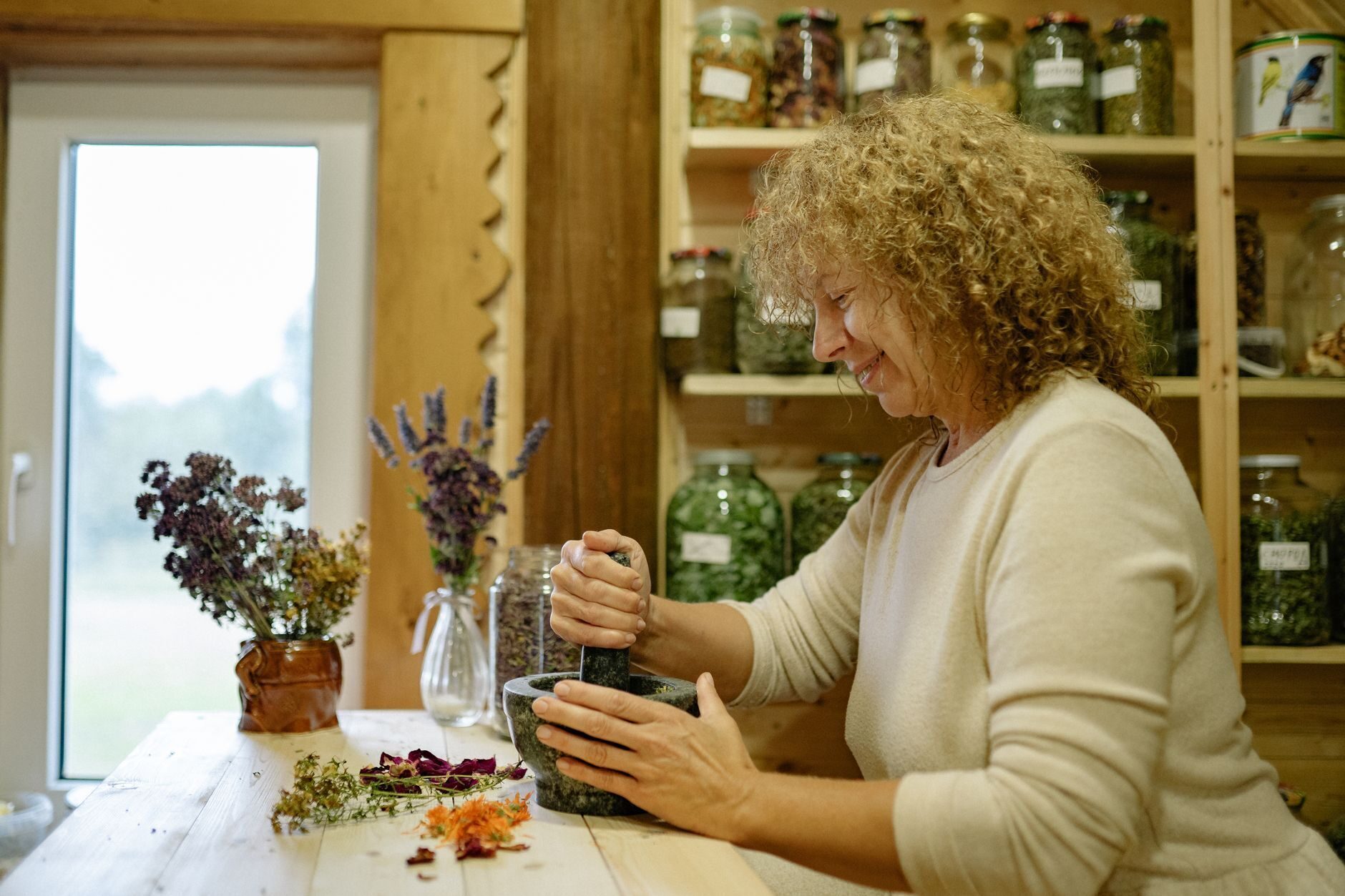 Herbalist in a cream sweater grinding herbs in a stone mortar before a wall of labeled glass jars