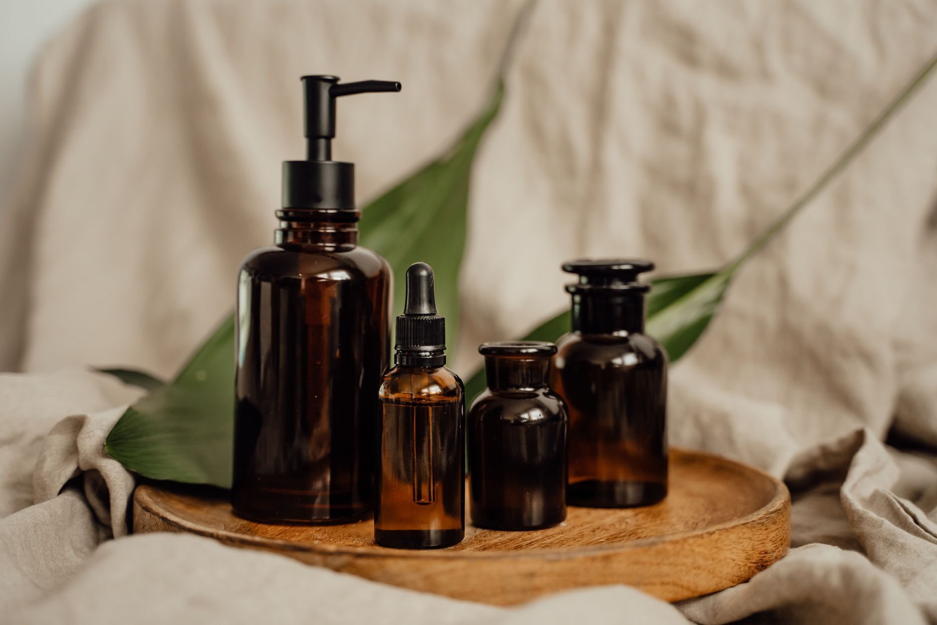 Amber glass dropper bottles arranged on a round wooden tray over warm linen with a single green leaf